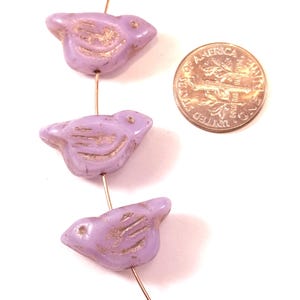 May include: Three light purple bird-shaped beads strung on a thin wire, featuring recessed wing details with a metallic gold finish. A United States dime is positioned next to the beads to provide a clear sense of scale for the items.