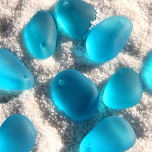 May include: A collection of smooth, light blue sea glass stones with holes drilled through them. The stones are arranged on a white sandy surface.