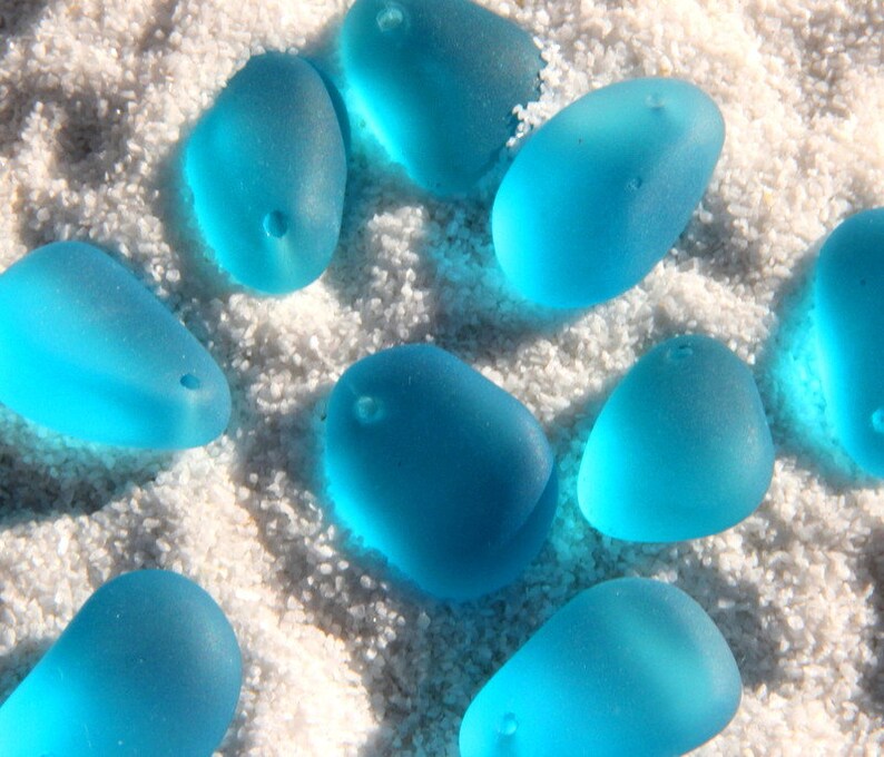 May include: A collection of smooth, light blue sea glass stones with holes drilled through them. The stones are arranged on a white sandy surface.