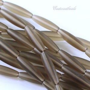 May include: Close-up of strands of elongated, oval-shaped beads in a muted brown color. The beads are strung together, creating a textured surface. The beads have a translucent quality, and the overall aesthetic is neutral and earthy.
