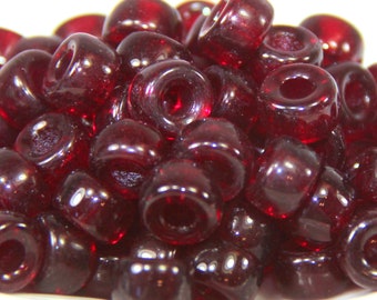 May include: A pile of translucent, dark red glass beads. Each bead is cylindrical with a large hole running through the center. The beads are smooth and glossy, reflecting light. These beads are suitable for jewelry making and crafts.