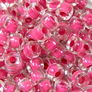 Pode incluir: Um close-up de um monte de contas de plástico rosa e transparentes. As contas são redondas e têm um furo no centro. São perfeitas para fazer joias ou outros trabalhos manuais.