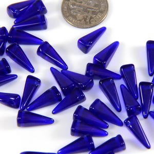 Könnte beinhalten: Eine Sammlung von kobaltblauen Glasperlen in Tropfenform mit einer spitzen Spitze. Die Perlen sind auf einer weißen Oberfläche verstreut.