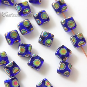 May include: A scattering of square glass beads with iridescent blue and green colors. The beads have a pattern of small circles on them.