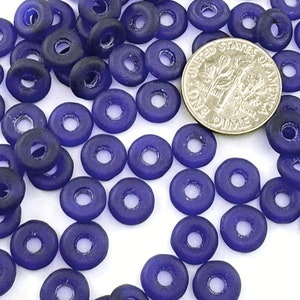 May include: A close-up of a pile of small, round, dark blue glass beads with a hole in the center. The beads are scattered on a white surface. A US dime is included for scale.