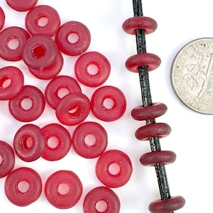 May include: A close-up of a pile of small, red glass donut beads. The beads are about the size of a US dime. The beads are strung on a black cord.