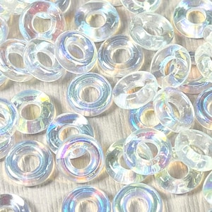 Puede incluir: Primer plano de numerosas cuentas transparentes iridiscentes en forma de anillo. Cada cuenta tiene un agujero central y muestra un espectro de colores. Ideal para la fabricación de joyas y proyectos de artesanía.