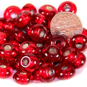 May include: A collection of translucent red glass beads with silver-colored centers and small white dots. A U.S. dime is included for size comparison. The beads are round with a hole through the center, suitable for jewelry making or crafts.