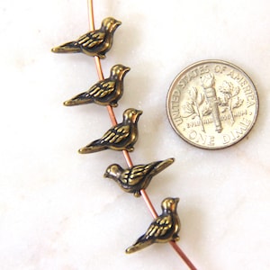 May include: Five small, antique brass-colored bird charms strung on a copper wire. The charms are detailed with etched feathers and are next to a U.S. dime for size comparison. The birds are facing left.