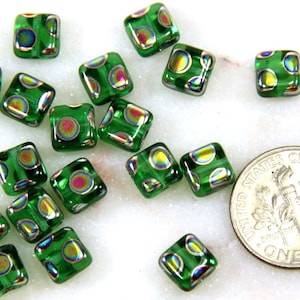 May include: A group of green square glass beads with iridescent circles. The beads are scattered on a white surface.