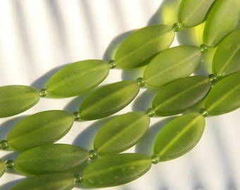 May include: Close-up of a necklace featuring olive-green, oval-shaped beads. The beads are strung together with smaller, round green beads. The necklace is displayed on a white surface, with shadows adding depth.