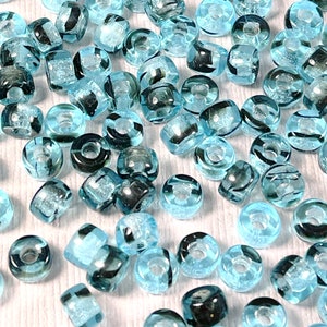 May include: A collection of translucent blue glass beads with dark gray accents. The beads are in various shapes, including round, square, and cylindrical, and are scattered across a white surface. The beads have a glossy finish and are suitable for jewelry making.