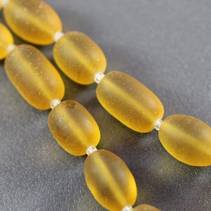 Puede incluir: Primer plano de un collar de cuentas ovaladas de color ámbar. Las cuentas tienen un aspecto esmerilado y están ensartadas con pequeñas cuentas transparentes. El fondo es de color gris oscuro sólido.