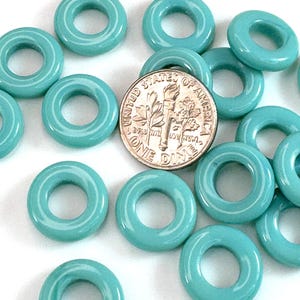 Puede incluir: Una colección de cuentas turquesas en forma de rosquilla. Se incluye una moneda de diez centavos de dólar estadounidense para comparar el tamaño. Las cuentas son brillantes y de color uniforme, con un agujero en el centro. Las cuentas son probablemente para hacer joyas o manualidades.