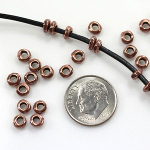 May include: A close-up of a pile of copper-toned metal beads, some of which are strung on a black leather cord. A US dime is included for scale.