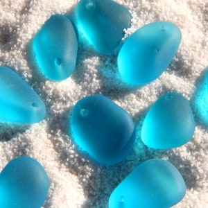 May include: A collection of smooth, light blue sea glass stones with holes drilled through them. The stones are arranged on a white sandy surface.