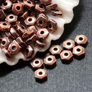 May include: A pile of copper-colored hexagonal beads with a hole in the center. The beads are arranged in a pile on a white seashell. There are also a few beads scattered on a dark gray surface.