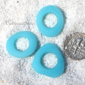 May include: Three turquoise blue sea glass rings with a hole in the centre. The rings are on a white sandy surface. Eastcoastbeads is written in the top left corner of the image.