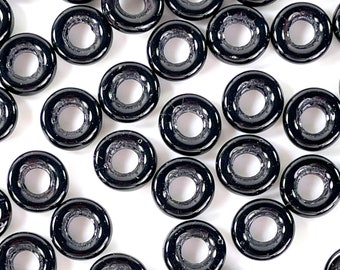 May include: A collection of glossy, black donut-shaped beads. Each bead has a circular hole in the center. The beads are scattered across a white surface, creating a pattern of light and shadow.