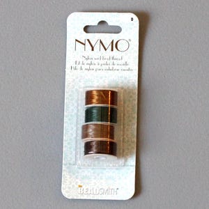 May include: A pack of NYMO nylon seed bead thread in brown, green and gold colours. The pack is labelled "NYMO Nylon seed bead thread" and "BEADSMITH".