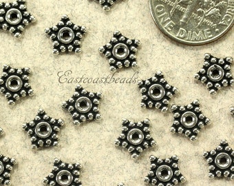May include: A close-up of a group of silver star-shaped beads with a small hole in the center. The beads are surrounded by a light brown surface.