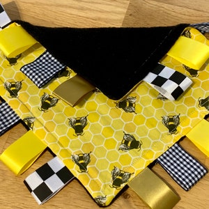 Puede incluir: Un juguete sensorial para bebés amarillo y negro con un patrón de panal y abejas. El juguete tiene múltiples cintas en blanco y negro a cuadros, amarillo y dorado.