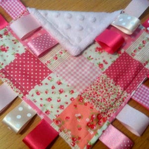 May include: A pink and white baby security blanket with a variety of patterned fabrics and ribbons. The blanket has a soft, plush texture and is perfect for babies to cuddle with.