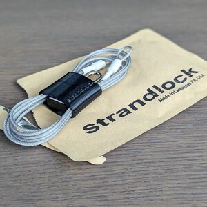 May include: A grey cable with a black magnetic cable organiser. The cable is coiled and the organiser is attached to the cable. The cable is packaged in a brown paper bag with the text "Strandlock Made in Lancaster PA USA".