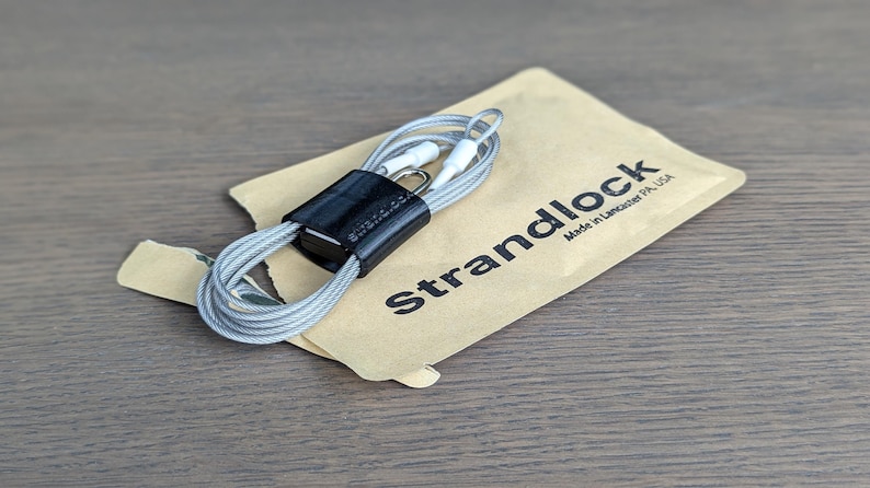 May include: A grey cable with a black magnetic cable organiser. The cable is coiled and the organiser is attached to the cable. The cable is packaged in a brown paper bag with the text "Strandlock Made in Lancaster PA USA".
