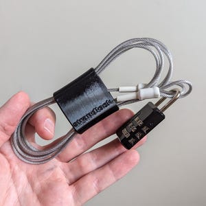 May include: A black combination lock with a silver cable attached to a black plastic cable organiser. The organiser has the word "strandtools" printed on it.