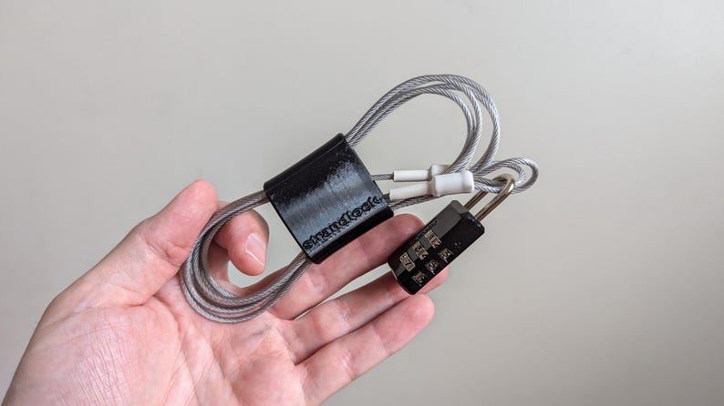May include: A black combination lock with a silver cable attached to a black plastic cable organiser. The organiser has the word "strandtools" printed on it.