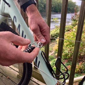 May include: A person secures a bicycle to a railing with a combination lock and cable. The bicycle is light green and has a black water bottle holder. The railing is light green and has a wooden deck behind it.