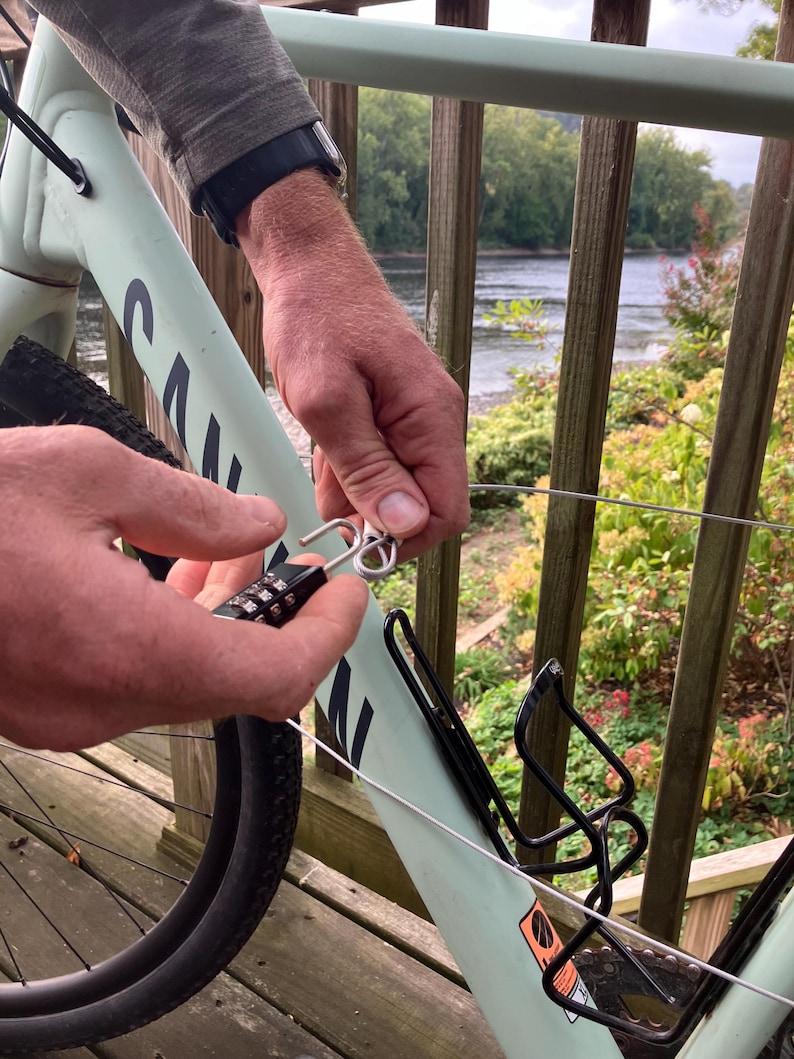 May include: A person secures a bicycle to a railing with a combination lock and cable. The bicycle is light green and has a black water bottle holder. The railing is light green and has a wooden deck behind it.