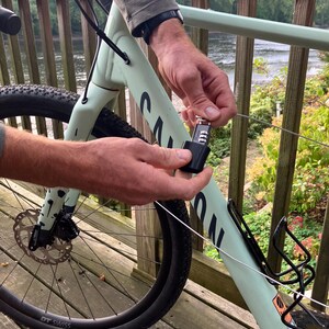 May include: A person secures a bicycle with a cable lock to a railing. The bicycle is light green with black accents and has a black front tire with a white "DT SWISS" logo. The cable lock is black and has a silver key.