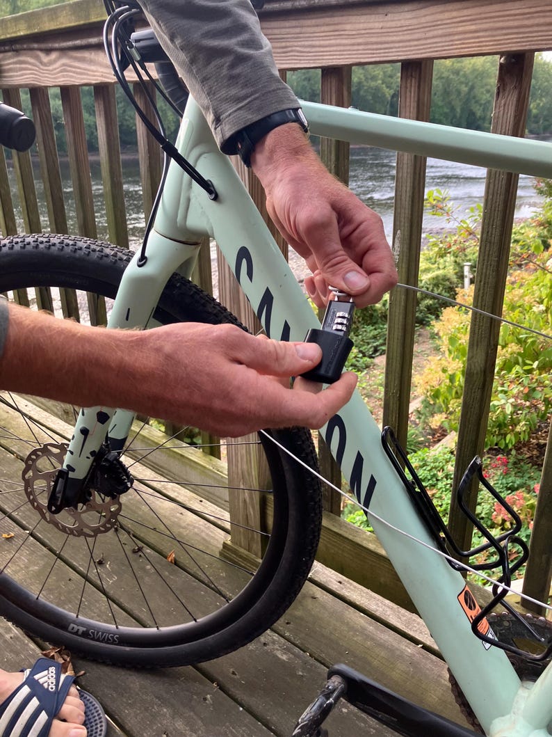 May include: A person secures a bicycle with a cable lock to a railing. The bicycle is light green with black accents and has a black front tire with a white "DT SWISS" logo. The cable lock is black and has a silver key.