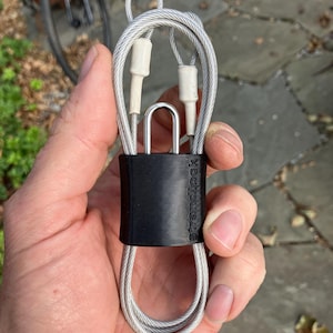 Puede incluir: Un candado de cable plateado con una funda protectora negra y la palabra "strandlock" impresa en él.