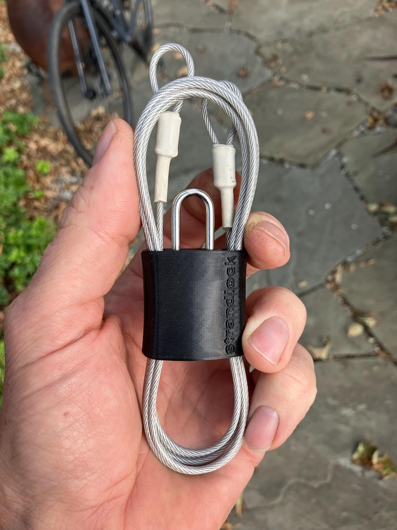 May include: A silver cable lock with a black protective sleeve and the word "strandlock" printed on it.
