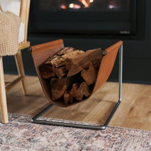 May include: A brown leather and metal firewood holder filled with logs. The holder has a rectangular metal frame and a curved leather sling to hold the wood. The firewood holder is placed on a wooden floor.