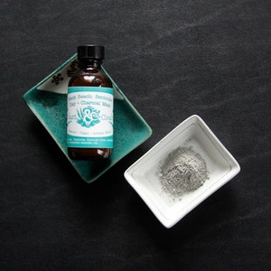 Pu&ograve; includere: Un flacone di vetro marrone con un'etichetta turchese che recita "Black Beauty Bentonite Clay + Charcoal Mask" e un logo verde e bianco. Il flacone &egrave; appoggiato su un piatto in ceramica turchese. Un piccolo piatto in ceramica bianco con polvere grigia all'interno &egrave; su una superficie nera.
