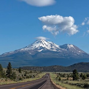 May include: A scenic view of a snow-capped mountain range with a winding road leading towards the peaks. The sky is a bright blue with fluffy white clouds.
