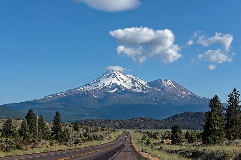 May include: A scenic view of a snow-capped mountain range with a winding road leading towards the peaks. The sky is a bright blue with fluffy white clouds.