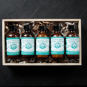 Pu&ograve; includere: Una scatola di legno piena di sei piccoli flaconi di vetro di maschera per il viso. Ogni flacone contiene una maschera di argilla diversa con un'etichetta che dice "Coast & Clearings". Le etichette sono blu turchese con testo bianco e un design a foglia verde.