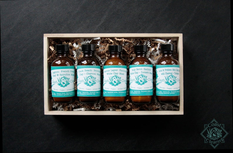 Pu&ograve; includere: Una scatola di legno piena di sei piccoli flaconi di vetro di maschera per il viso. Ogni flacone contiene una maschera di argilla diversa con un'etichetta che dice "Coast & Clearings". Le etichette sono blu turchese con testo bianco e un design a foglia verde.