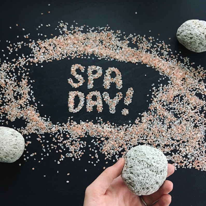 May include: A black background with pink Himalayan salt scattered around the words "SPA DAY!" written in white salt. Three gray pumice stones are also shown, one in a hand.