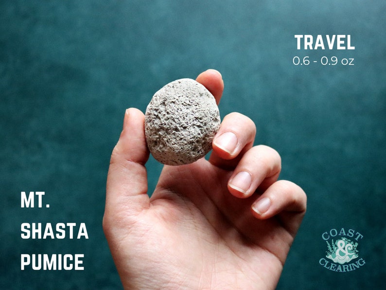 May include: A hand holding a small, gray pumice stone. The stone is labeled "MT. SHASTA PUMICE" and "TRAVEL 0.6 - 0.9 oz".