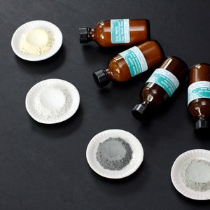 Pu&ograve; includere: Cinque piccoli flaconi di vetro con etichette e un design verde turchese. Ogni flacone &egrave; accanto a un piccolo piatto bianco contenente una polvere di colore diverso: giallo, bianco, grigio e verde.