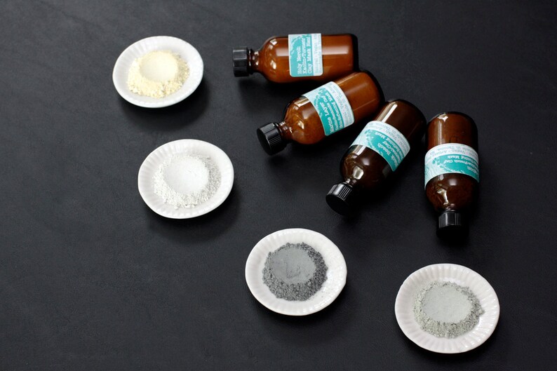 Pu&ograve; includere: Cinque piccoli flaconi di vetro con etichette e un design verde turchese. Ogni flacone &egrave; accanto a un piccolo piatto bianco contenente una polvere di colore diverso: giallo, bianco, grigio e verde.