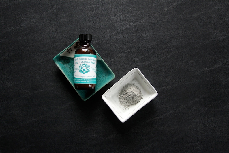 Pu&ograve; includere: Un flacone di vetro marrone con un'etichetta turchese che recita "Black Beauty Bentonite Clay + Charcoal Mask" e un logo verde e bianco. Il flacone &egrave; appoggiato su un piatto in ceramica turchese. Un piccolo piatto in ceramica bianco con polvere grigia all'interno &egrave; su una superficie nera.