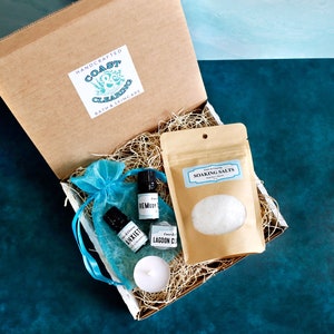May include: A brown cardboard box filled with bath and skincare products. The box contains a small bag of soaking salts, two small bottles of essential oils labeled "Anxiety" and "Remember", a teal blue organza bag, and a white candle. The box is labeled "Handcrafted Coast Clearing Bath & Skincare".