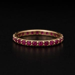 May include: A gold eternity band featuring a continuous row of small, round, vibrant red rubies set in a shared-prong setting. The ring has a polished metal finish and a uniform circular shape, showcasing a series of gemstones around the band.